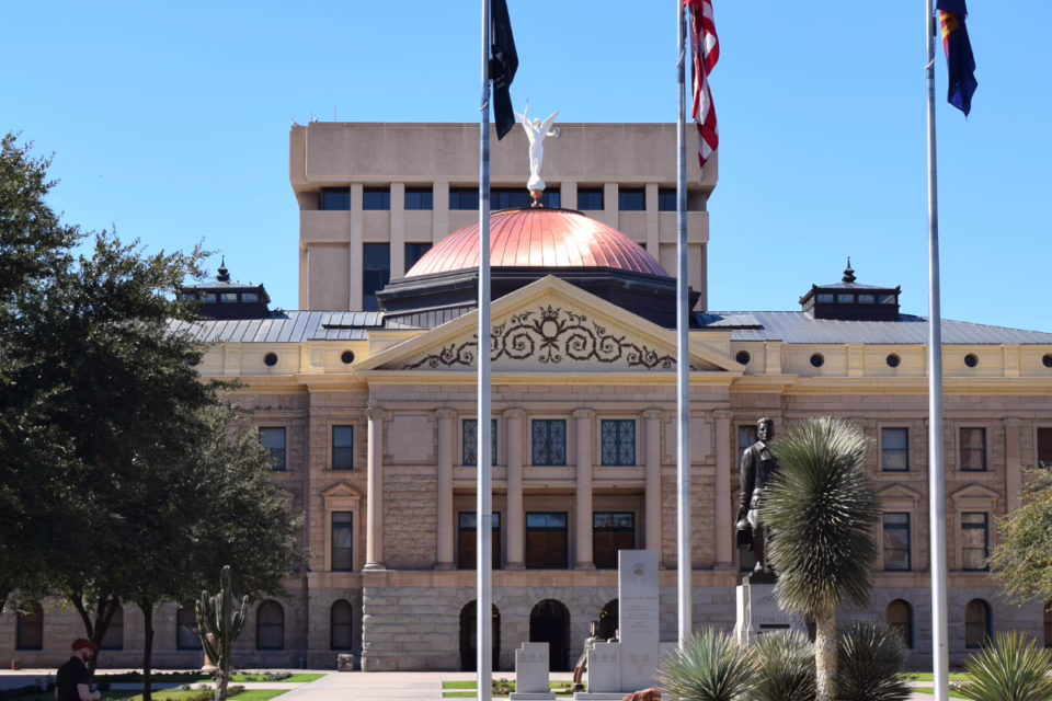 Arizona State Capitol with flags and a statue in front, under a clear blue sky