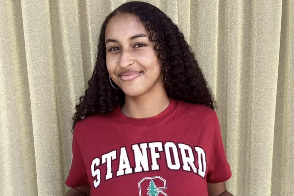 Student in a red Stanford shirt smiles in front of a beige curtain