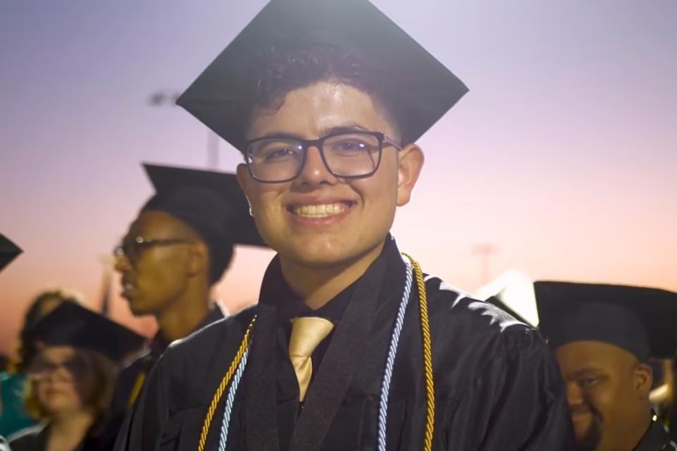 Graduate in cap and gown smiling at sunset, surrounded by fellow graduates