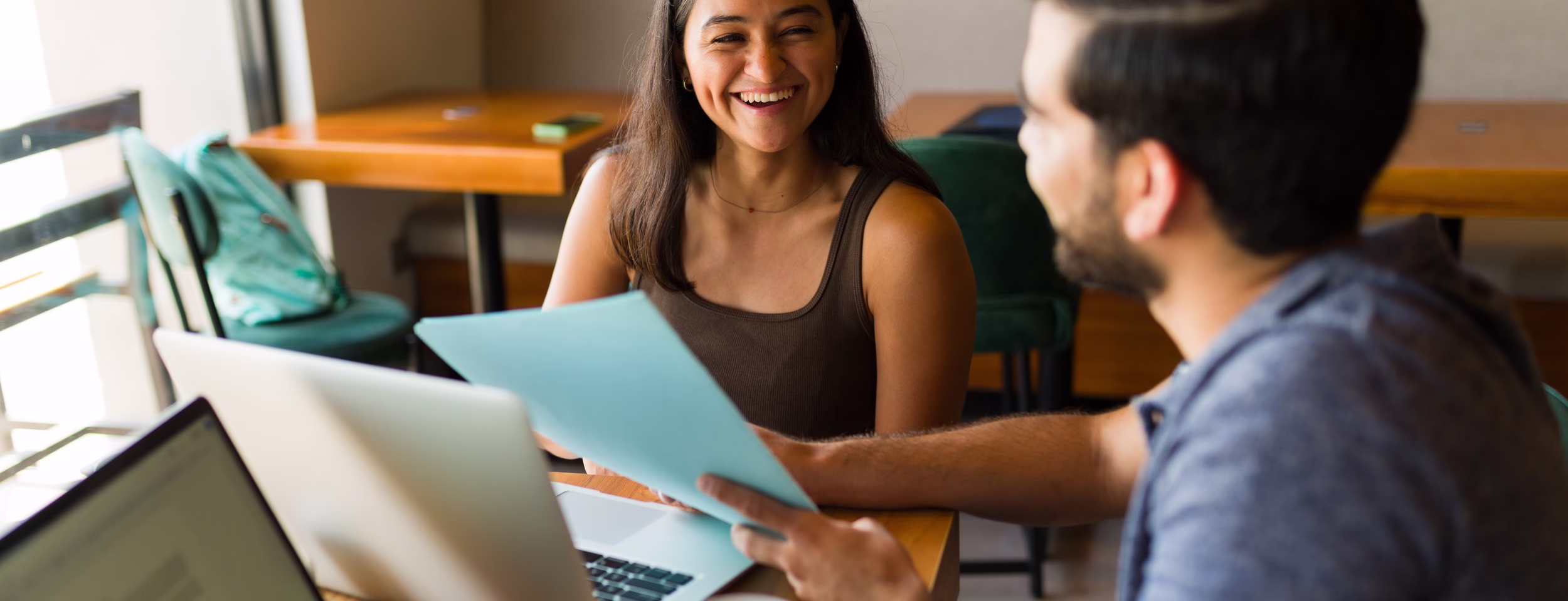 Two people smiling and discussing documents at a table with laptops and a coffee cup