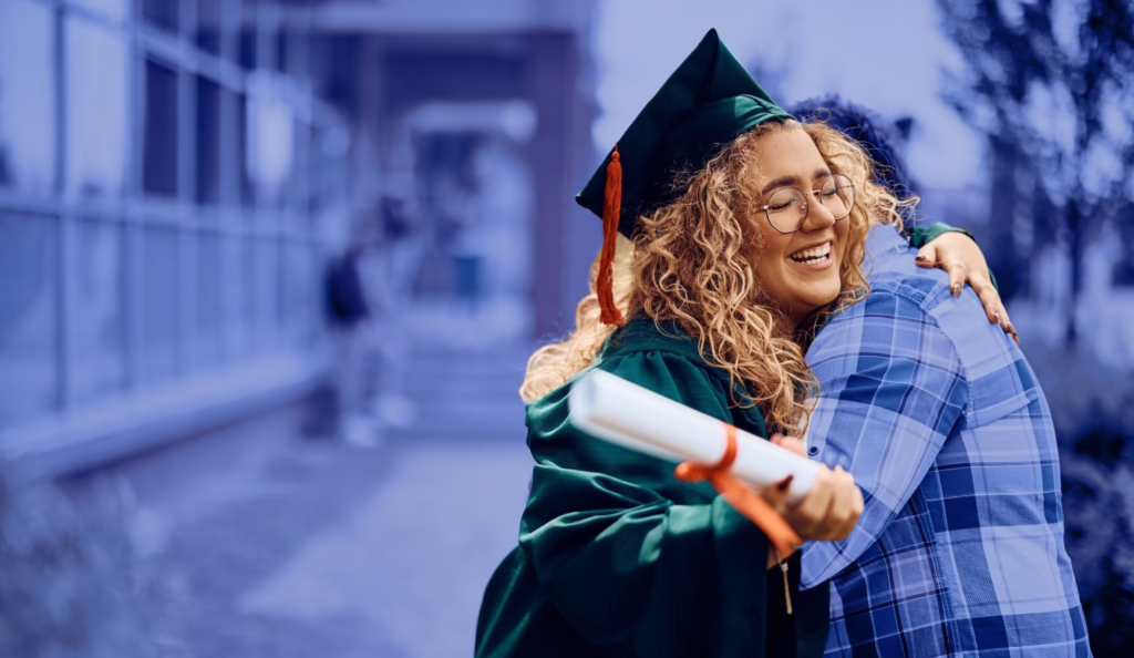 Graduate in cap and gown hugging someone, holding a diploma, smiling