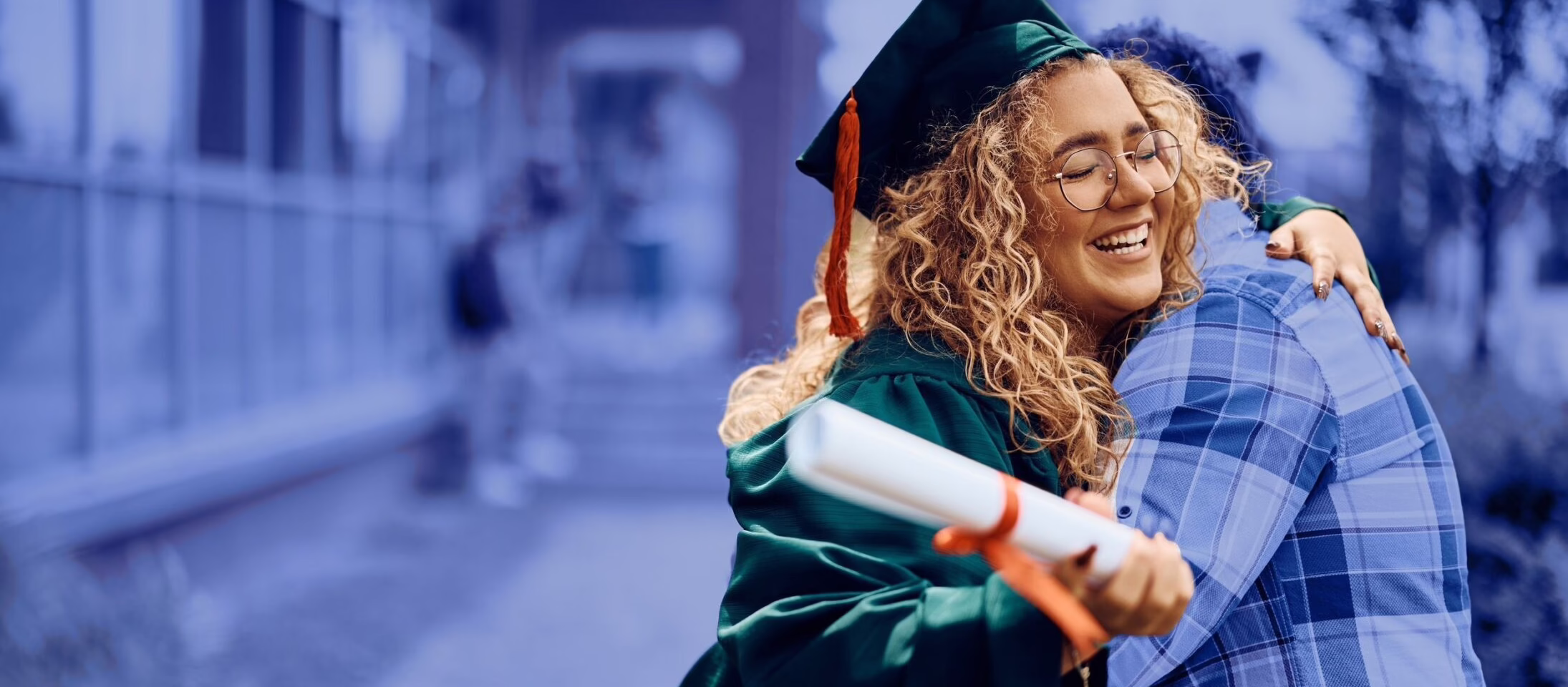 Graduate in cap and gown hugging someone, holding a diploma, smiling