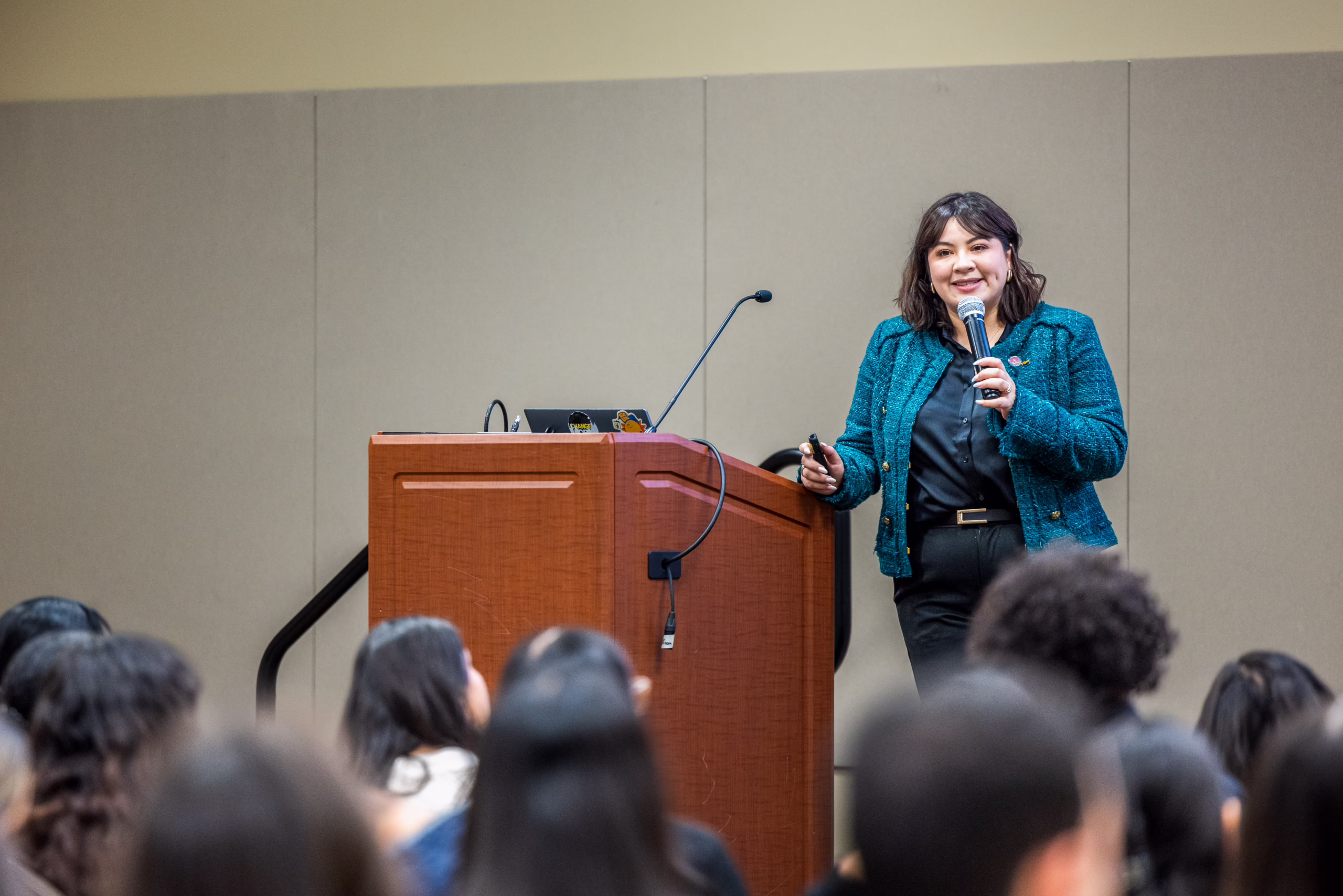 Speaker in a teal jacket presenting with a microphone at the Annual Leadership Symposium, audience in foreground